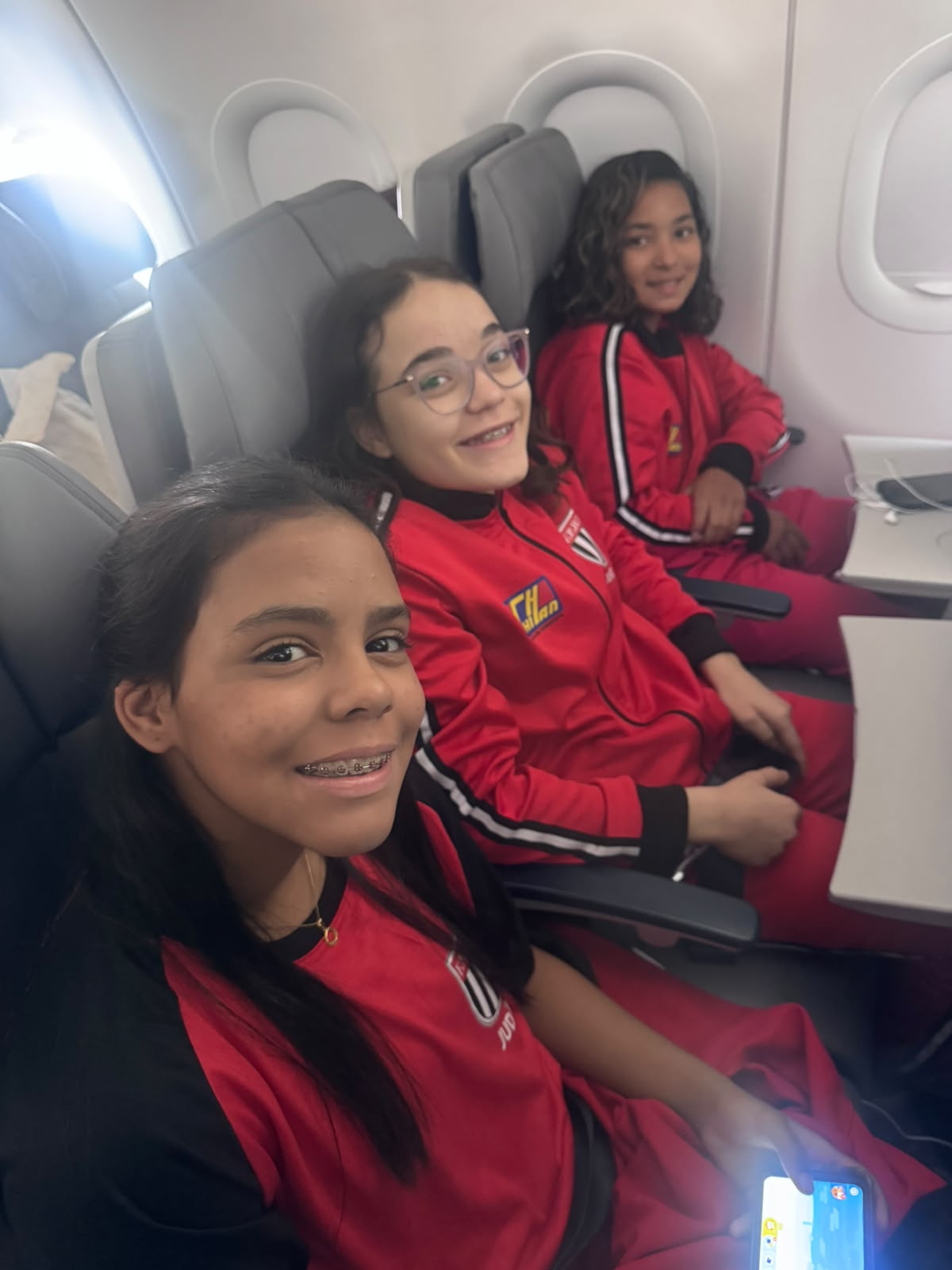 Lídia Sayuri e duas companheiras da seleção paulista de judô sentadas no avião, de agasalho vermelho