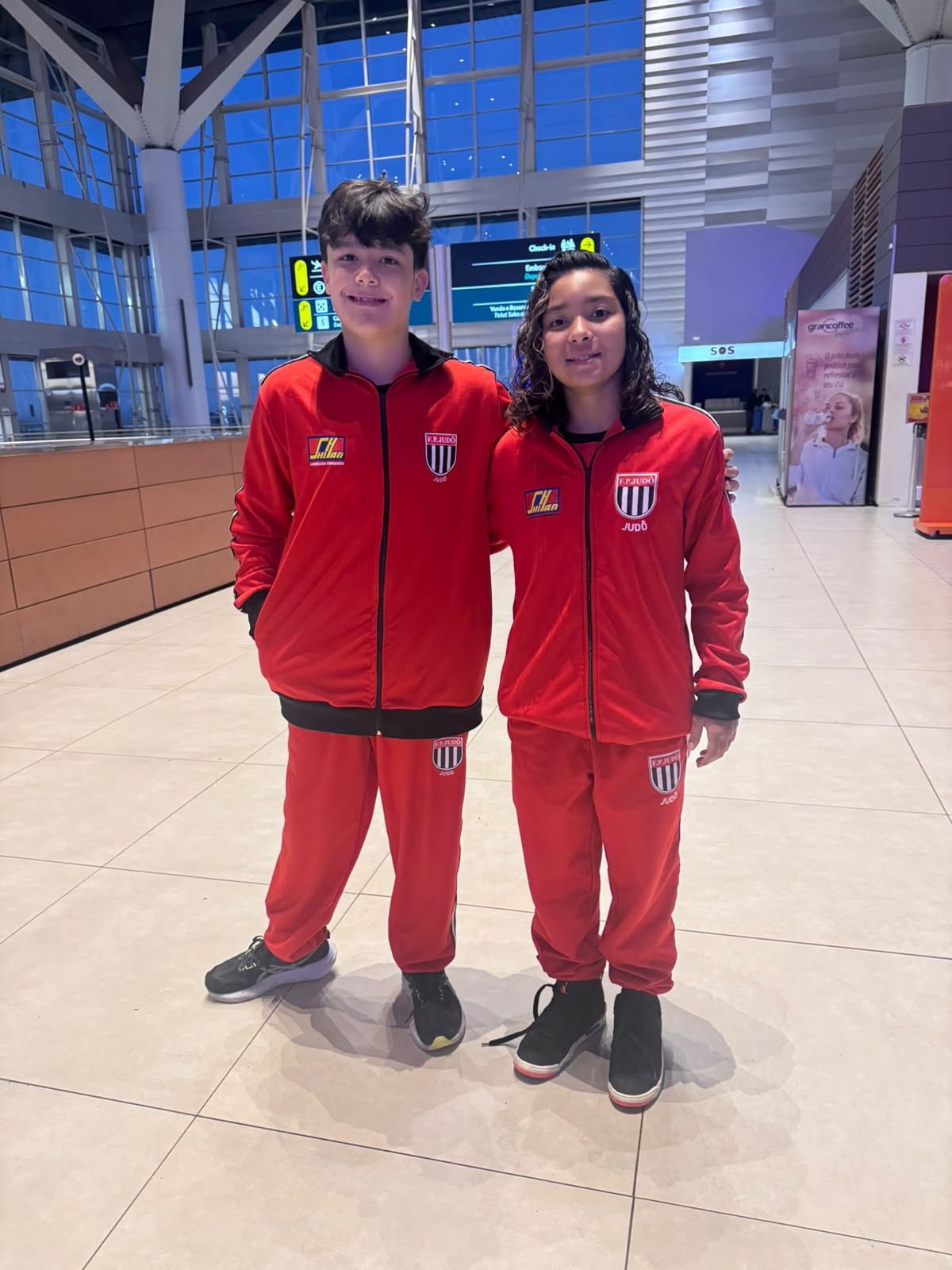Davi Von Ah Camargo e Lídia Sayuri Tsuji Palma lado a lado, de agasalho vermelho da seleção paulista de judô, no saguão do aeroporto