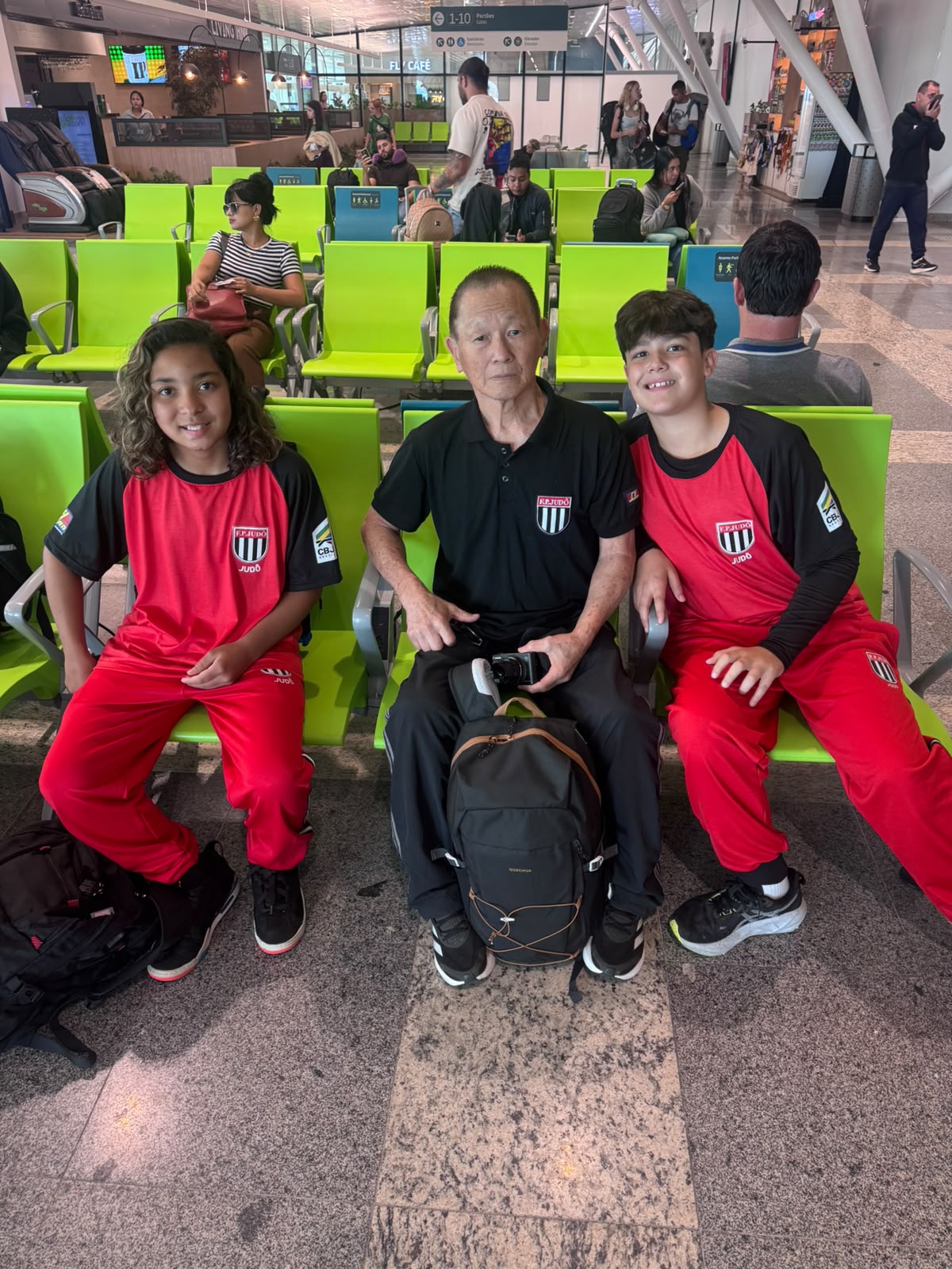 Lídia Sayuri, Sensei Sergio Honda e Davi Von Ah sentados na área de embarque do aeroporto, de agasalho da seleção paulista