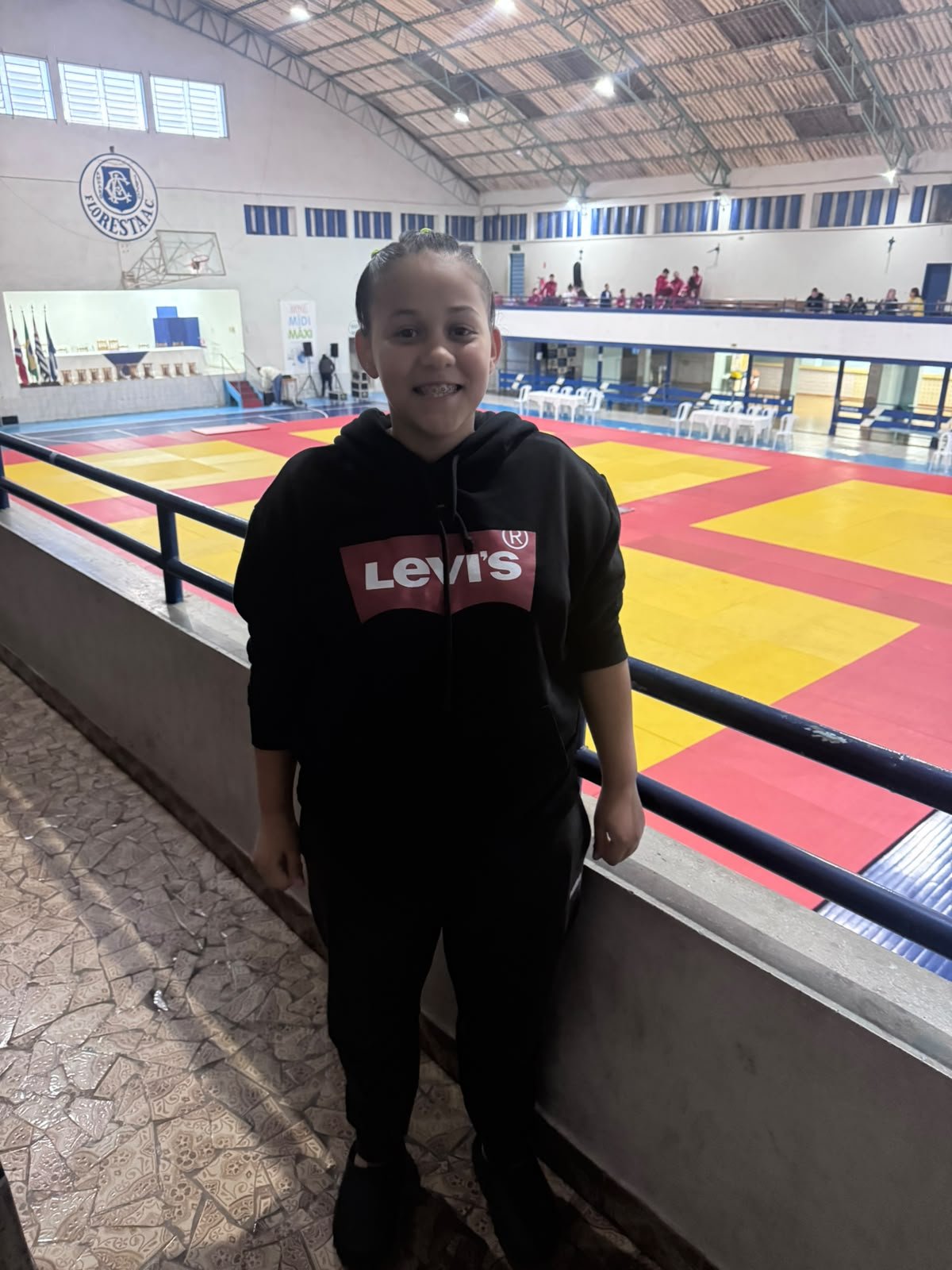 Clara na arquibancada do ginásio da Copa Amparo 2026, com os tatames ao fundo