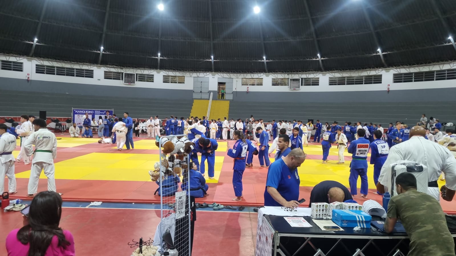 Vista panorâmica do Ginásio Domingão em Aguaí à noite, cheio de atletas em atividade, com o painel dos convidados olímpicos ao fundo