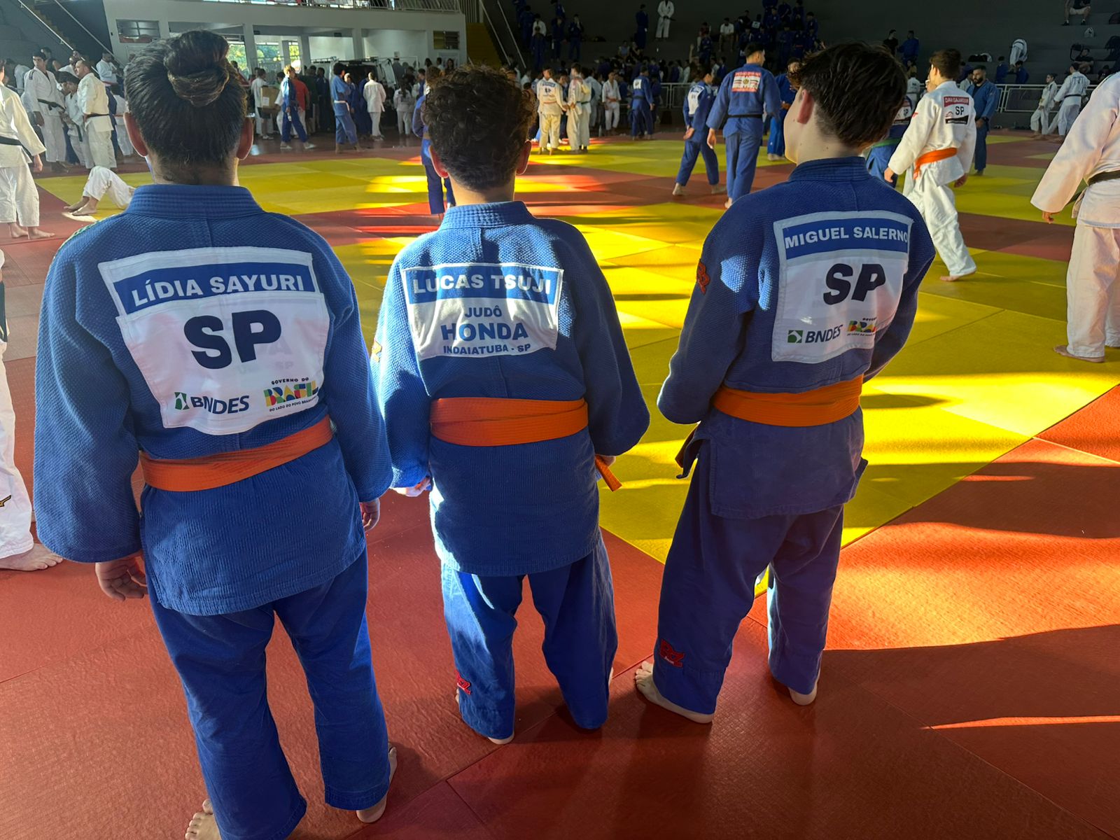 Trio Judô Honda de costas no tatame do Ginásio Domingão em Aguaí: Lídia Sayuri com dorsal SP da Federação Paulista, Lucas Tsuji com kimono Judô Honda Indaiatuba e Miguel Salerno com dorsal SP