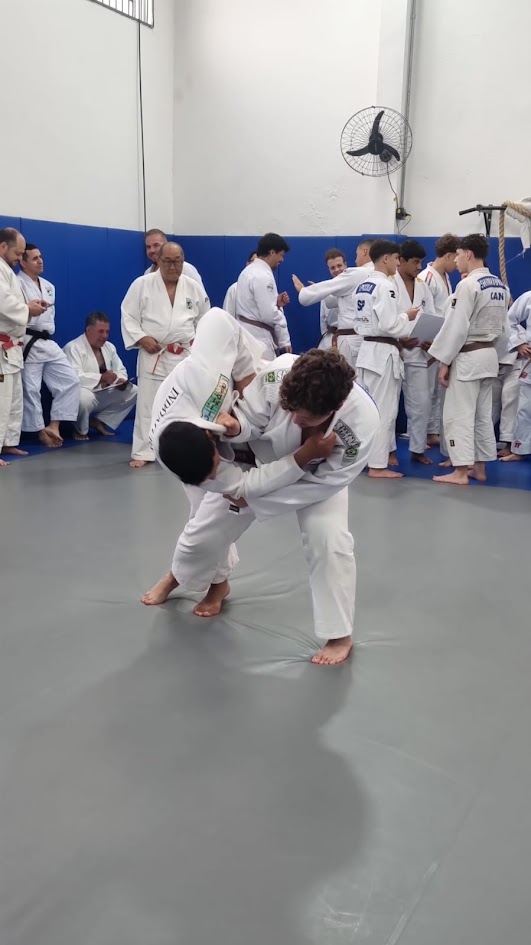 Vinícius Akira Pinto entrando um golpe durante o Módulo de Fundamentos Técnicos, com uke à sua frente e outros candidatos observando ao fundo