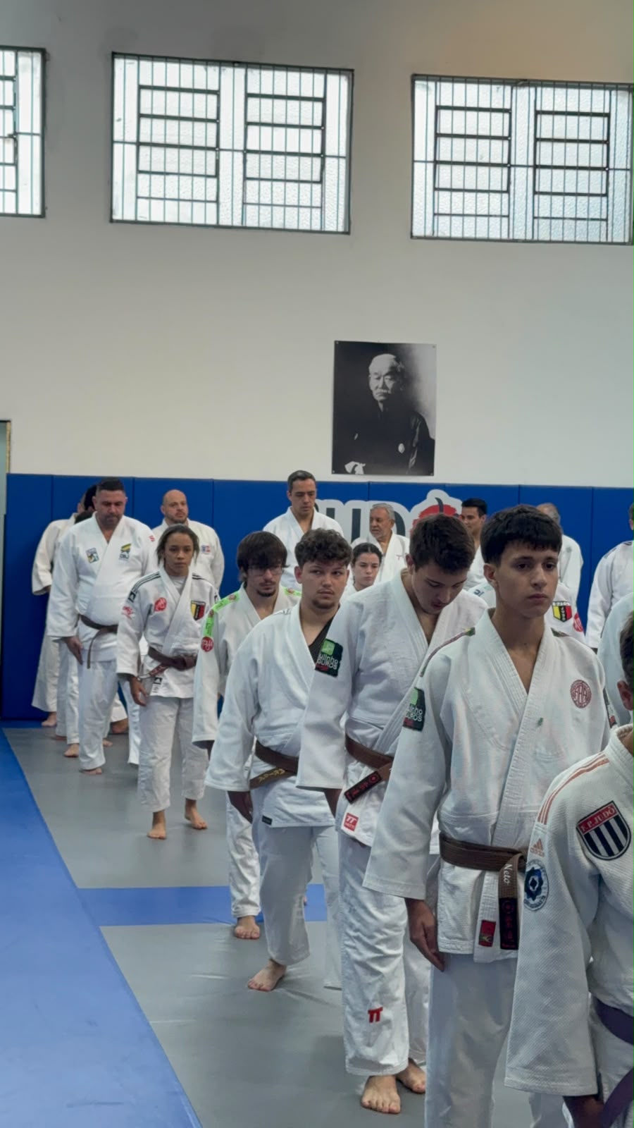 Cumprimento formal com atletas em fila voltados para o retrato de Jigoro Kano na parede da Academia Yamamoto
