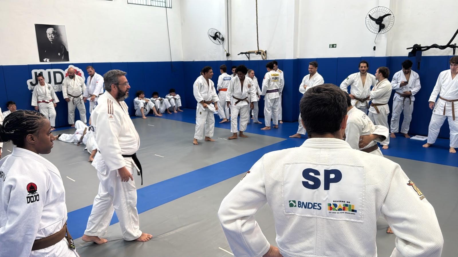 Tatame da Academia Judô Yamamoto durante o segundo dia do Módulo de Nage-no-Kata, com candidatos e senseis reunidos e o retrato de Jigoro Kano na parede