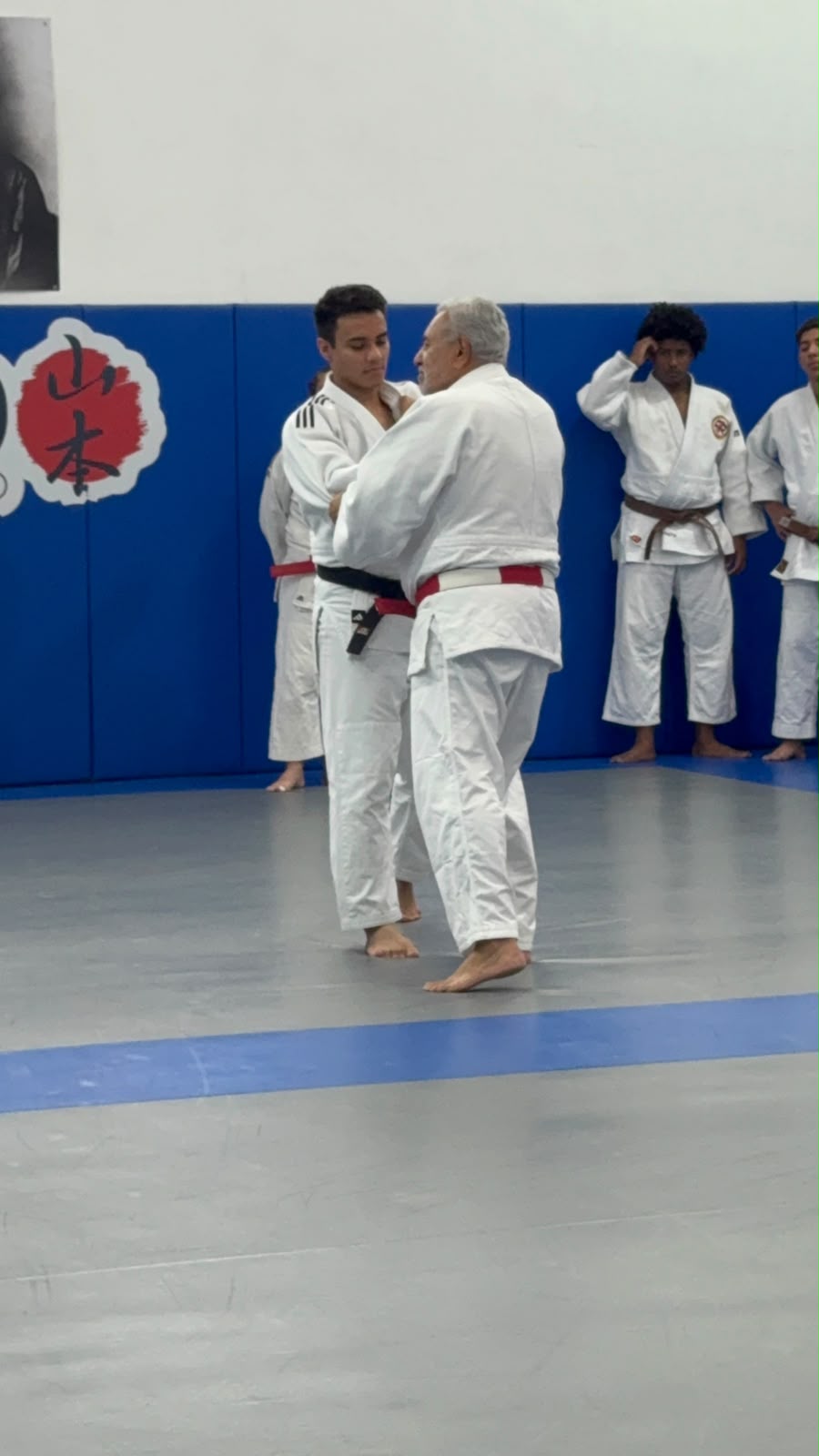 Sensei de faixa vermelha corrigindo pegada com Vinícius (uke) no tatame