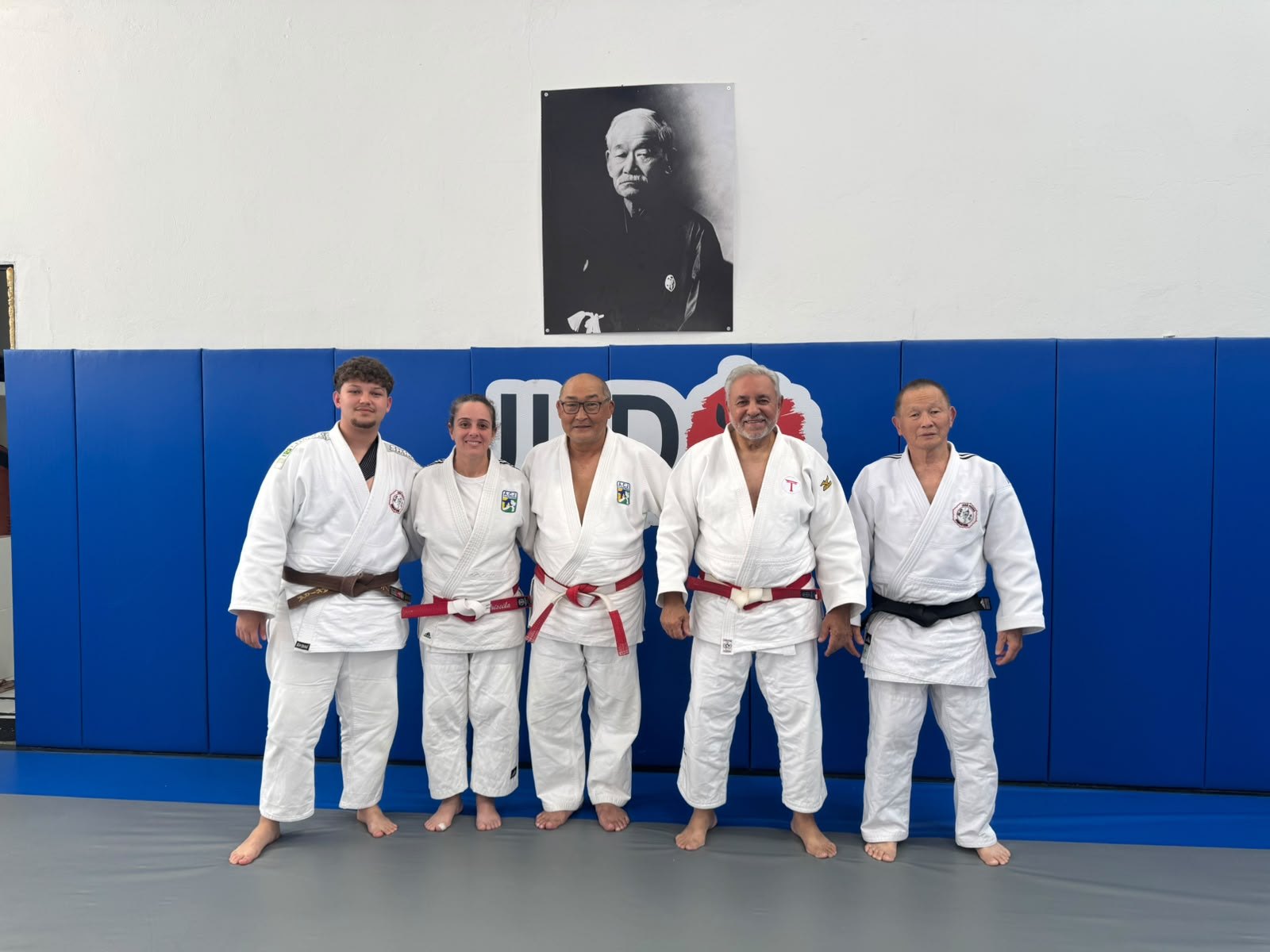 Vinícius Akira Pinto ao lado de três senseis de faixa coral e do Sensei Sergio Honda, sob o retrato de Jigoro Kano na Academia Judô Yamamoto