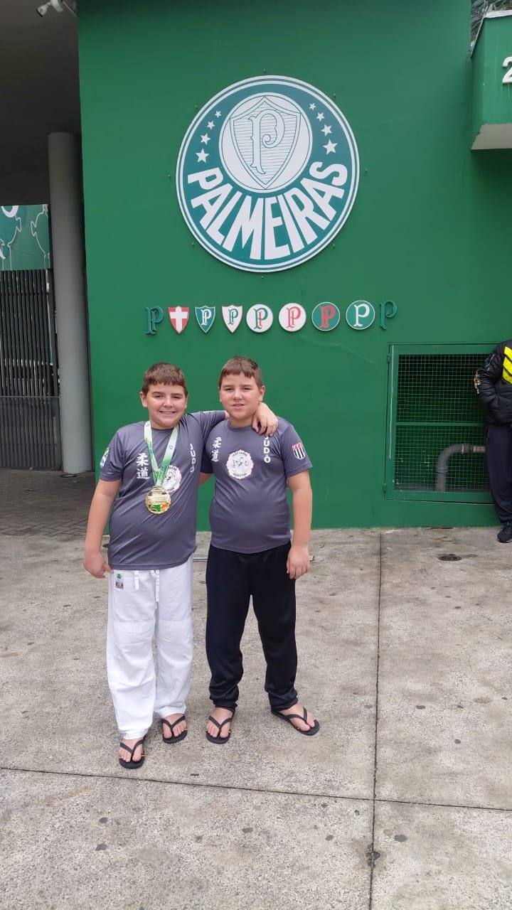 Heitor e Lorenzzo Pereira Lopes da Silva em frente ao escudo do Palmeiras, na entrada do Ginásio Palestra Itália, um deles com a medalha de ouro no peito
