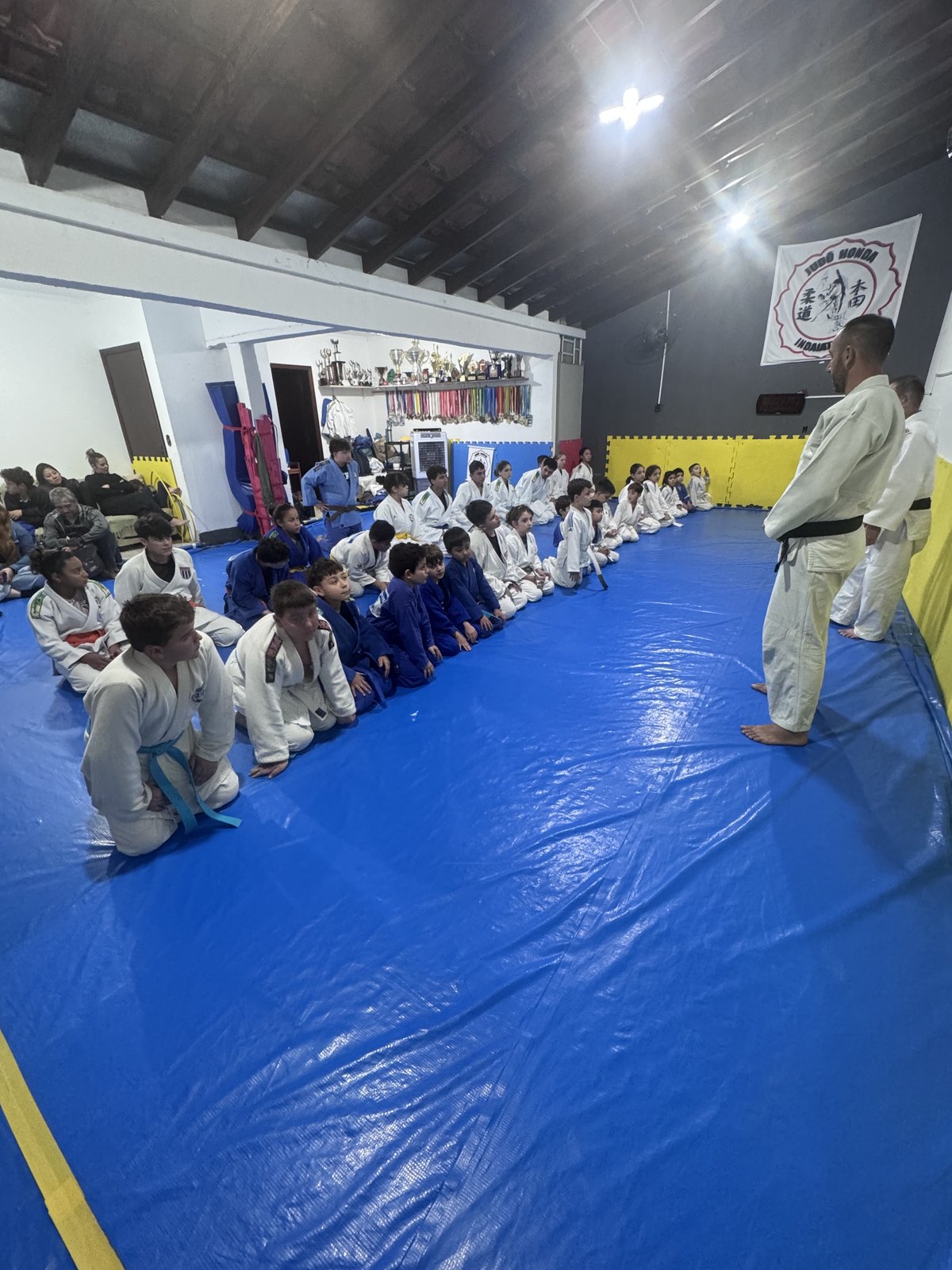 Tatame do Judô Honda cheio de alunos de judogi branco e azul durante o treino, com a estante de troféus do dojô ao fundo