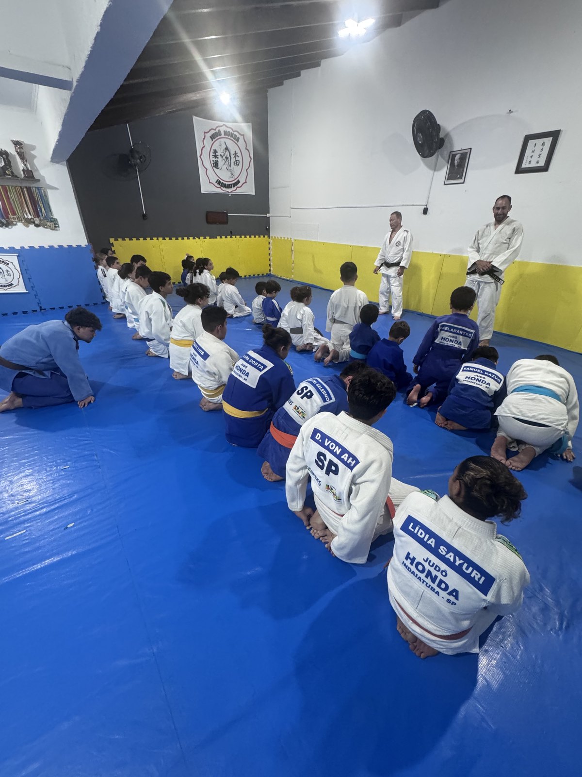 Alunos do Judô Honda sentados no tatame de costas, com os dorsais de competição com os nomes Lídia Sayuri e D. Von Ah